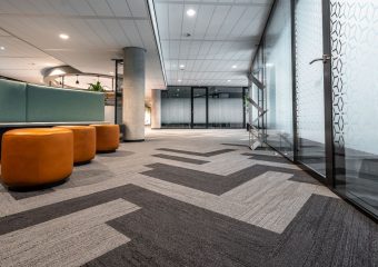Spacious and modern office corridor featuring glass doors, geometric carpet patterns, and leather seating.