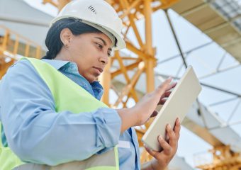 A female engineer in workwear checks her tablet on a construction site, highlighting modern engineering practices.